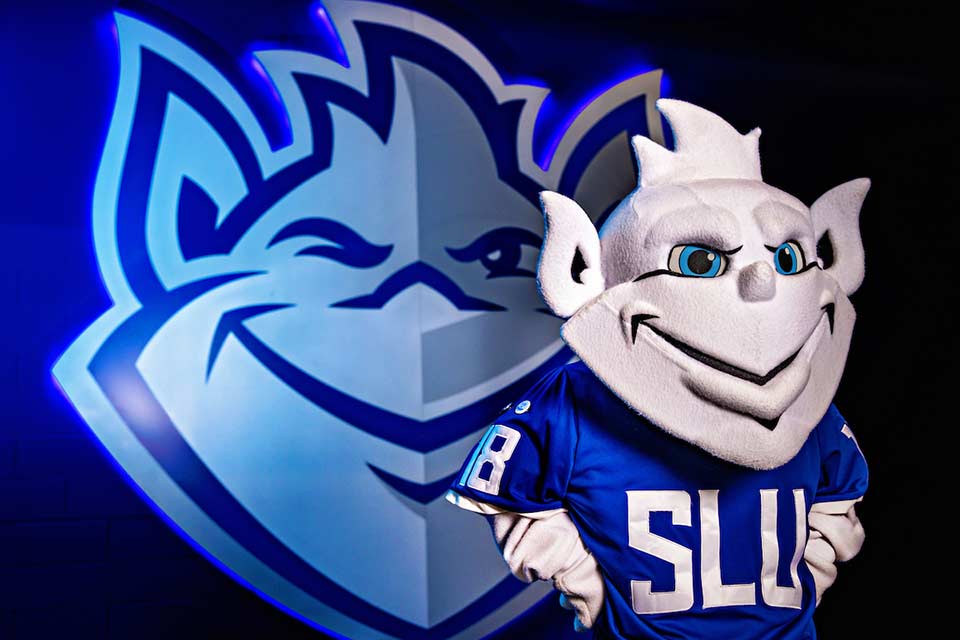 Billiken Mascot The Billiken mascot poses in front of the Billiken logo.