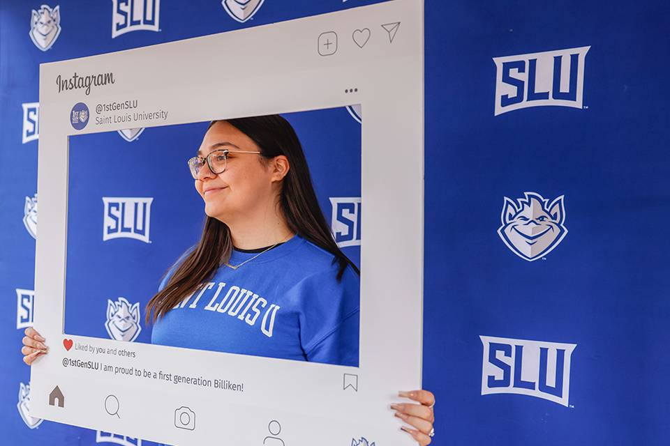 First Generation A student wearing a SLU sweatshirt stands in front of a SLU banner while holding up a picture frame that says First Generation at SLU and Proud to be a first-generation Billiken