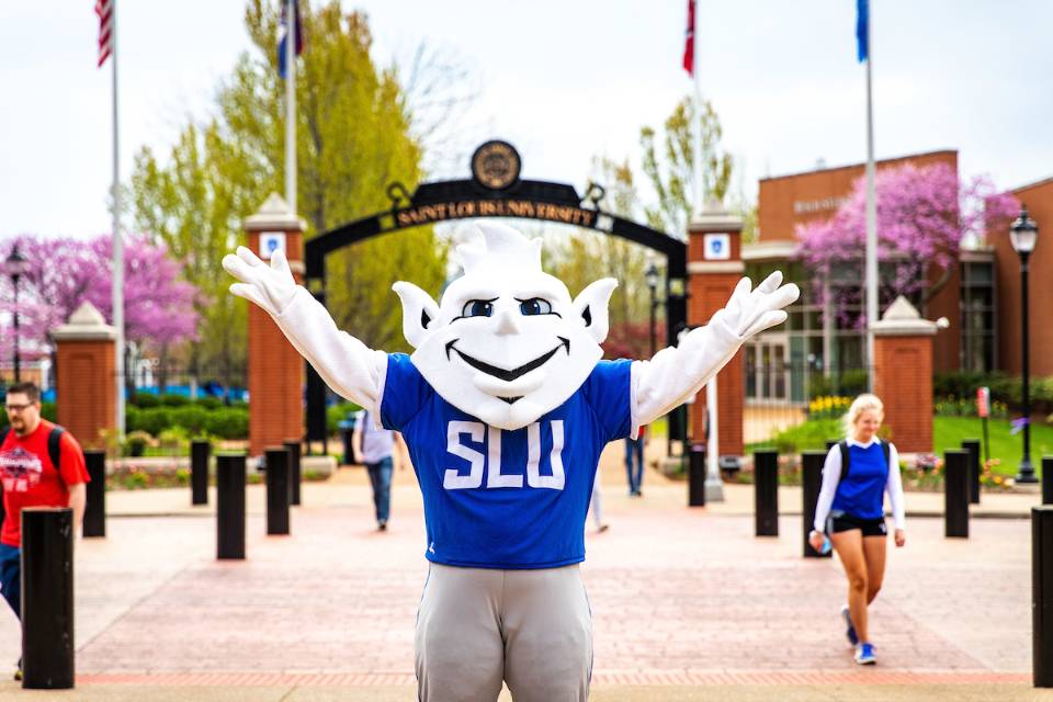 The Billiken poses for a photo outside on campus. Students cross the street behind him.