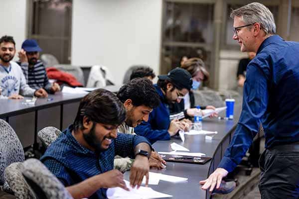 Students sit and work on an assignment in a lecture-hall style classroom. A male faculty member stands in front of them.