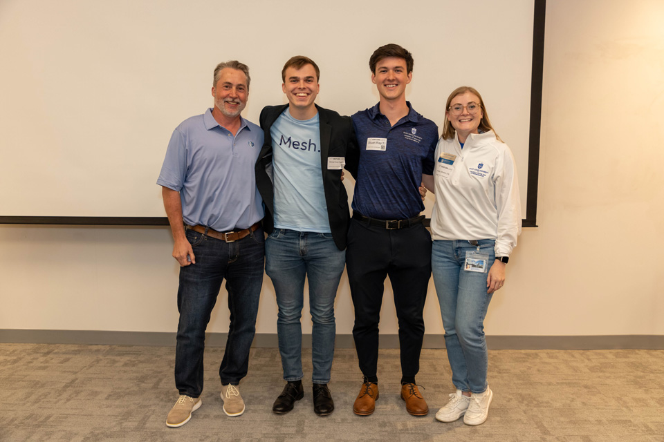 Four students, faculty and staff stand at the front of a classroom in front of a whiteboard.