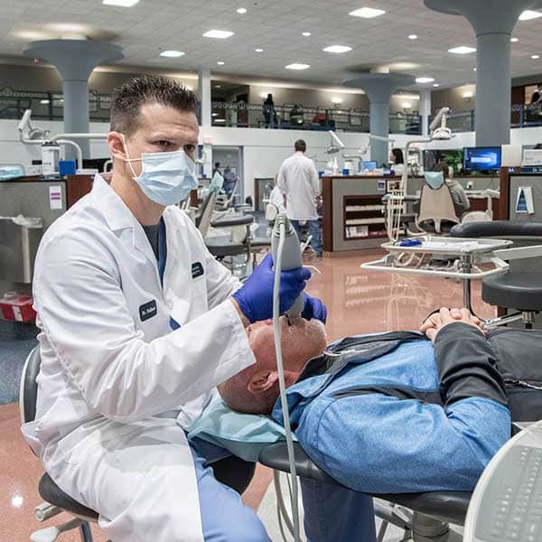 A male CADE resident, wearing a white coat and a medical mask, holds a medical device to the mouth of a reclining patient in a dental chair. The dentist is looking at a screen while he works.