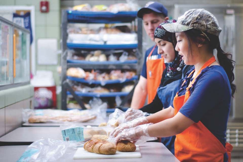 Students prepare meals at SLU's Campus Kitchen Three students are shown in profile, wearing aprons, working with bread on a kitchen counter.