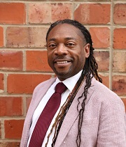 Richard Marks headshot Richard Marks, wearing a pink suit, maroon tie and white shirt, stands in front of a brick wall and smiles