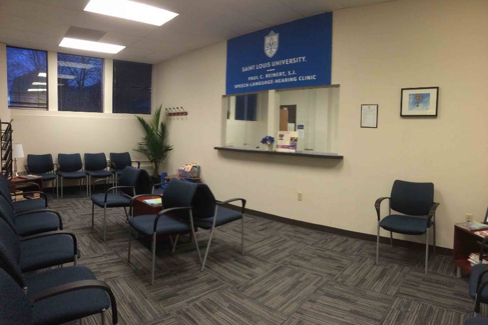 Clinic interior Interior view of a waiting room with chairs and plants. A large sign with the SLU logo and the words "Paul C. Reinert Speech Language and Hearing Clinic" hangs above the reception window.