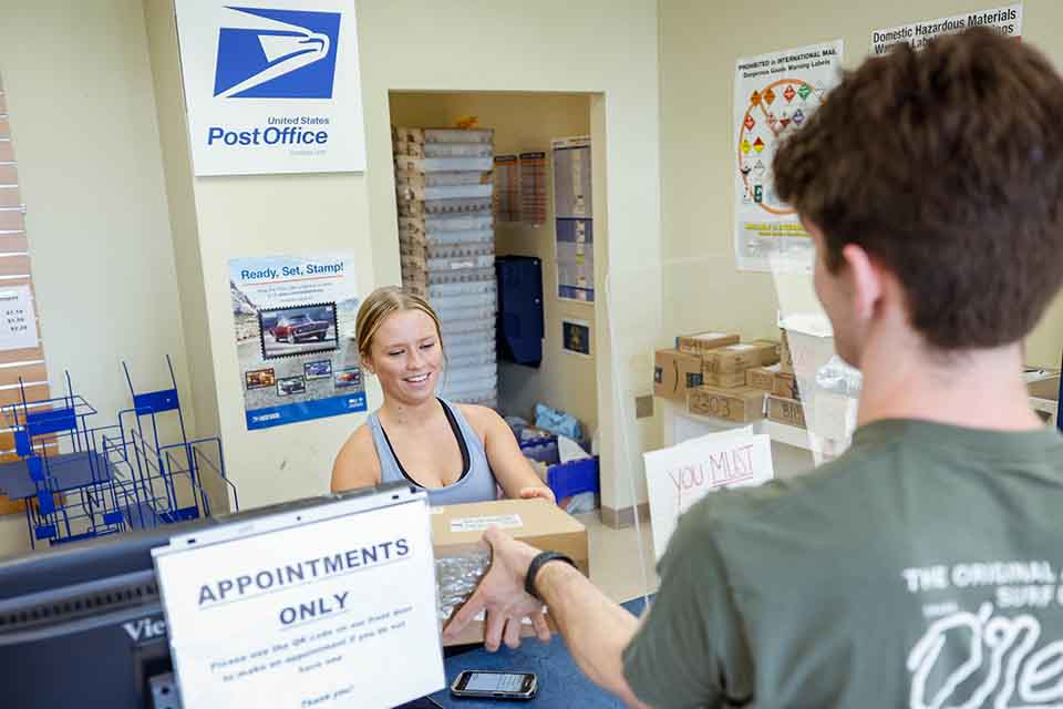 Inside the SLU mailroom A mailroom worker surrounded by packages hands a student (seen from the back) his mail