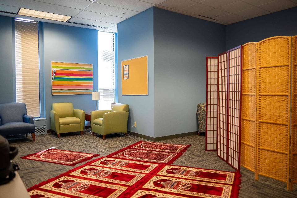 B S C Interfaith space. A room with blue walls and comfortable chairs. Prayer rugs are seen lined up on the floor.