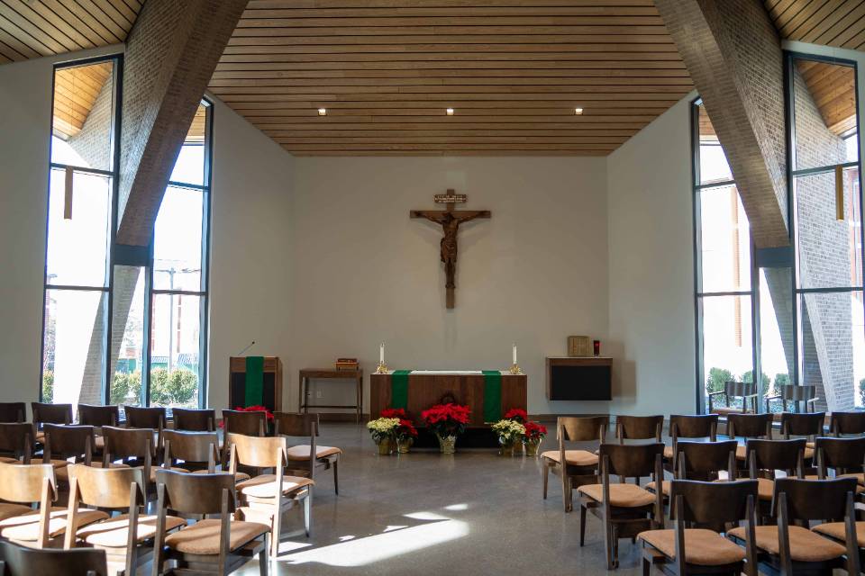 Jesuit Center View of a chapel with rows of chairs seen from the back, with an aisle down the middle. At the front, there is an altar, crucifix. Red and white poinsettias are on display. There are tall windows on each side, with a slatted wooden ceiling.