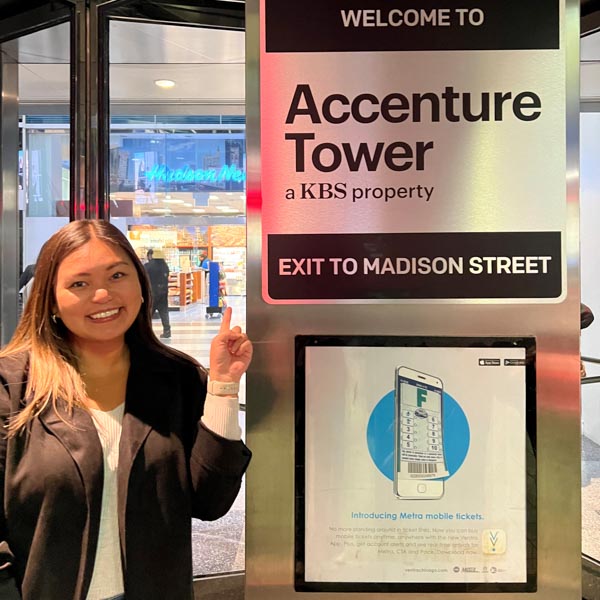 Daniella Soriano standing to the left of Accenture Tower sign. She is smiling and pointing at the sign. The full sign reads "Welcome to Accenture Tower, a KBS property. Exit to Madison Street."