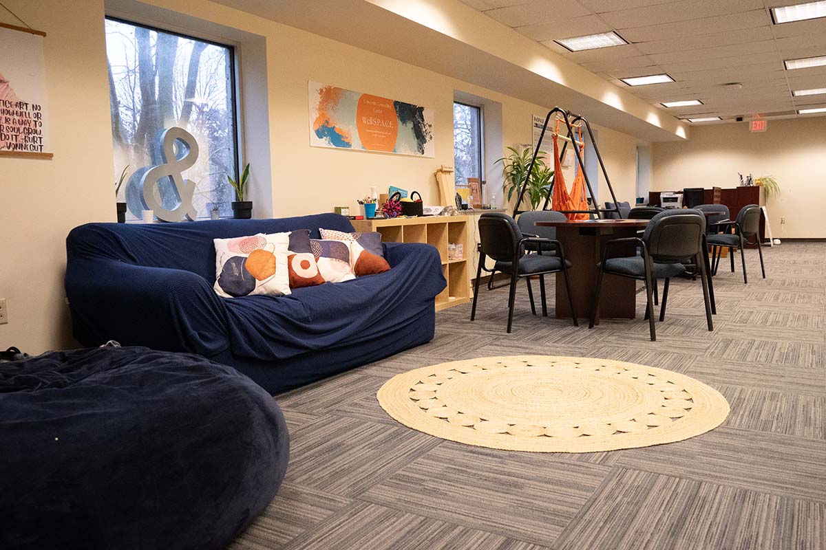 Wuller Hall WellSPACE Image of a room with carpeting, a round area rug, a comfy looking couch, bean bag chair, and tables with chairs.