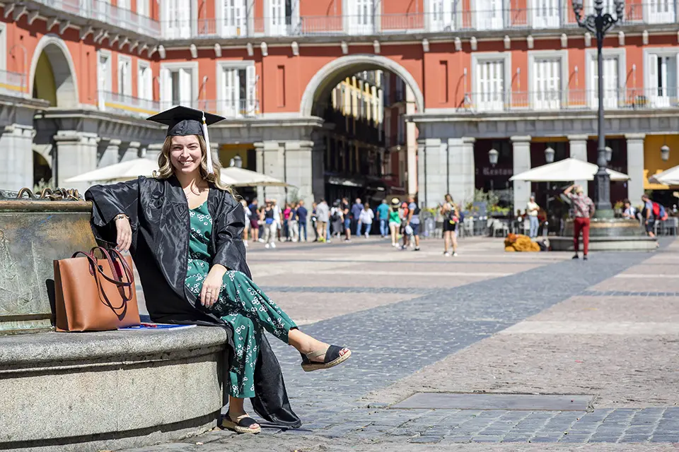 Honors Program student Honors Program student in graduation cap and gown with honors cords sits on a bench in surrounded by brick buildings in Madrid's Plaza Mayor.