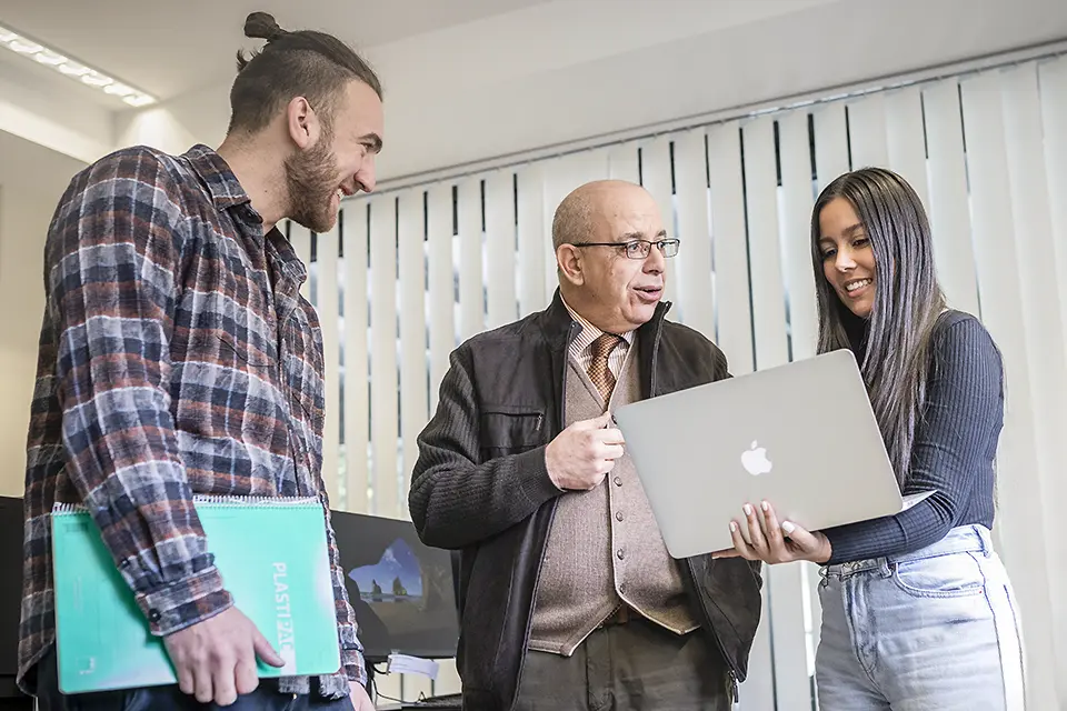 Man talking with two students while one of them is holding a laptop. 