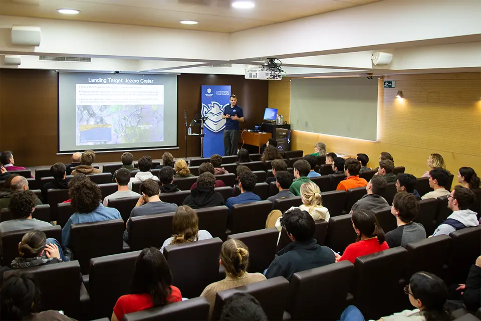 A man on stage speaks to audience in auditorium with a science presentation projected on the screen. 