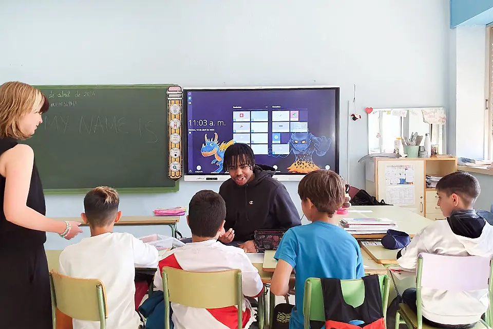 A SLU student sits talks to a group of four elementary students, seen from behind, sitting at desks in a classroom.