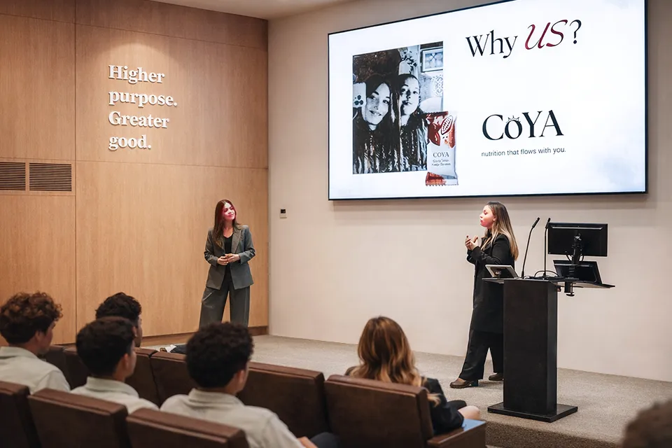 Camila Gallardo and Emilie Sarmiento present COYA in Viña Aula 1 Two women in business attire speak to the audience with a presentation slide that shows the company logo. The wall of the auditorium has a sign that reads "Higher purpose. Greater good" in white.