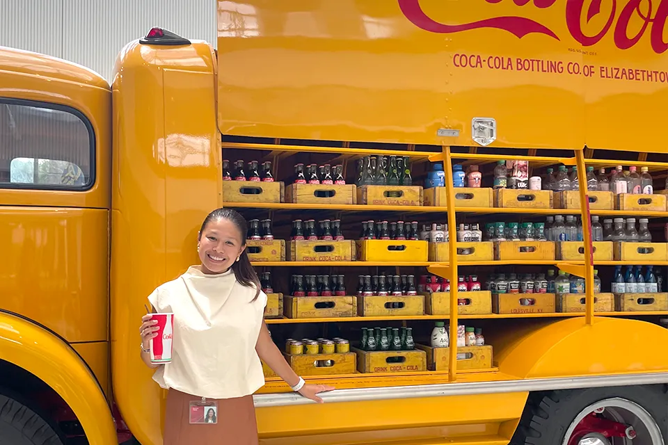 Reagan Tran, Learning Operations Intern at the global Coca-Cola headquarters. A young girl standing in front of a yellow truck holding a glass of soda in her hand.