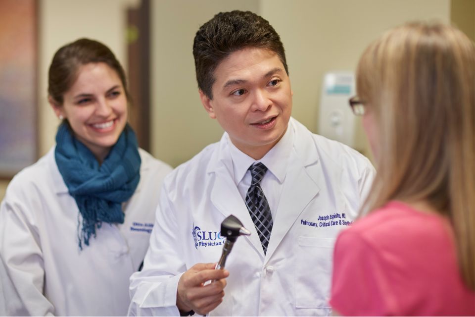 Two pulmonologists, one holding an otoscope, talk with a patient inside a hospital exam room.