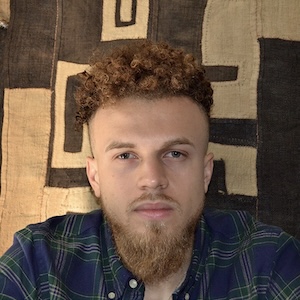 Bentley Brown A shoulder-length photograph of an adult man with auburn hair in tight curls, close cropped along the sides, a moustache and a well-trimmed, triangle-shaped beard, gazes calmly straight ahead. He wears a blue and green flannel shirt and sits in front of a brown and beige swath of fabric with an abstract design.