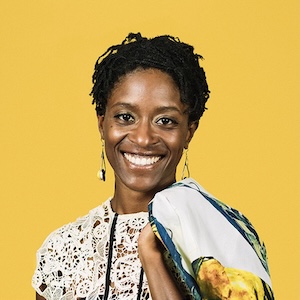 Dail Chambers A shoulder-length photograph of an adult woman with tightly braided black hair wearing dangling earrings and a floral-lace pattern top, holding a floral-print wrap draped across her left shoulder. She stands in front of a canary yellow background and smiles warmly.