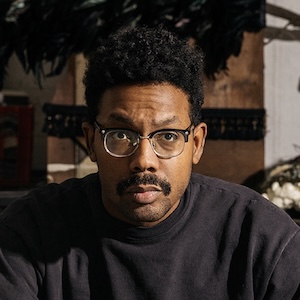 Damon Davis A shoulder-length photograph of a man with textured black hair, a moustache, and wearing black-rimmed glasses. His face is dramatically illuminated from the left and he looks intently straight ahead, his right eyebrow slightly raised. He wears a black crew-neck shirt. The room in the background is out of focus.