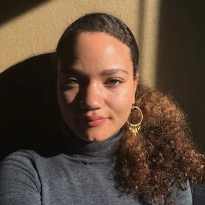 Summer Sloane-Britt A shoulder-length photograph of a young adult woman with auburn-tinted black hair pulled back in a pony tail that drapes across her left shoulder. She wears a heather-gray turtleneck sweater and large gold hoop earrings. She stands against a caramel-colored background. Golden light dramatically illuminates the left side of her face and leaves the right side in shadow.