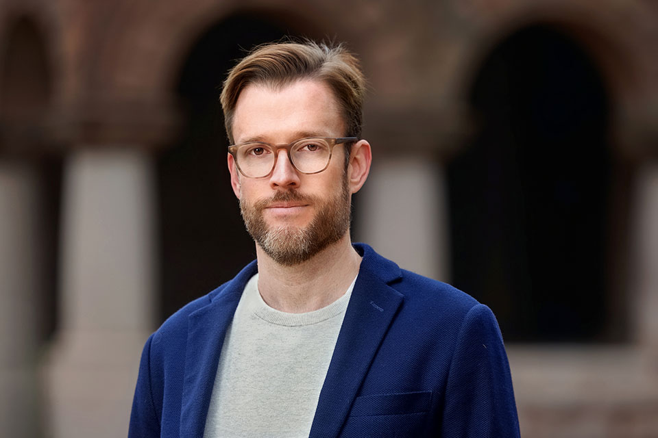 Bruce O’Neill, a man with glasses and a beard, stands confidently outside, wearing a blue blazer and gray shirt. Background features blurred stone arches.