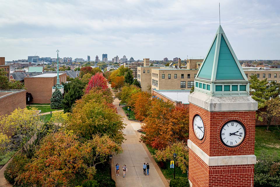 Aerial view of a college campus in autumn, featuring a brick clock tower with teal accents. Colorful trees line the walkway with distant city skyline. Aerial view of a college campus in autumn, featuring a brick clock tower with teal accents. Colorful trees line the walkway with distant city skyline.