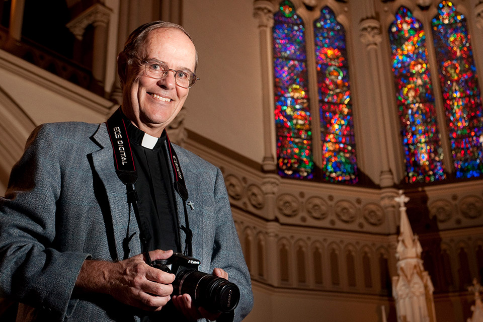 JJ Mueller with camera near stained glass An older white man wearing a Roman collar and a camera around his neck stands near stained glass windows.