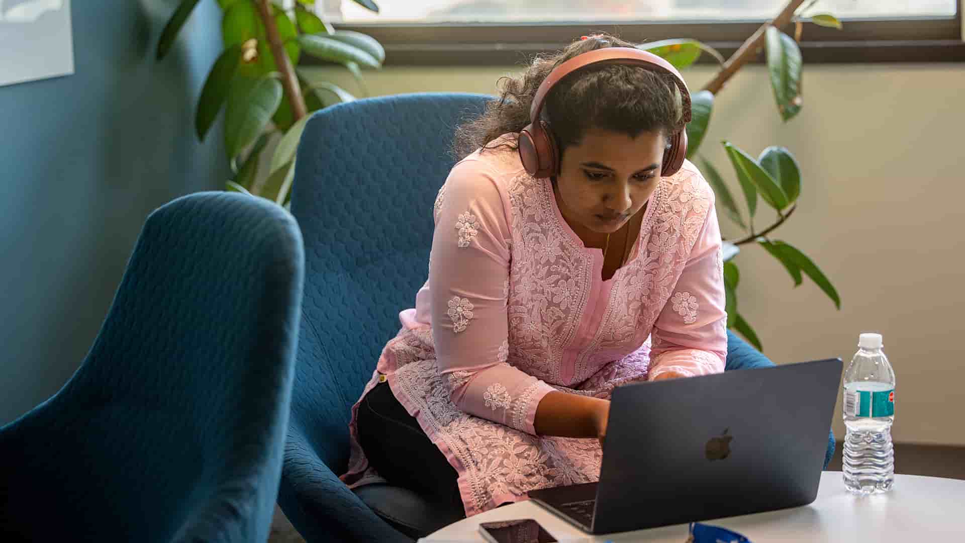 A student studies in a lounge space with cozy green chairs and plants along the wall of windows.