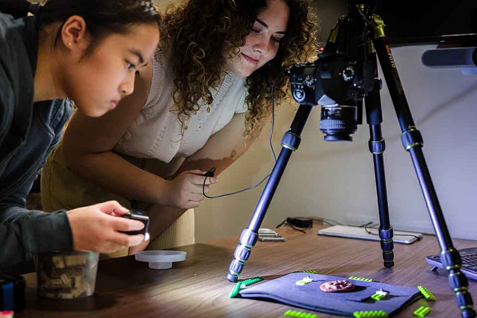 Two students lean over a table to examine a round, copper-colored artifact that is placed under a camera-like device attached to a tripod.