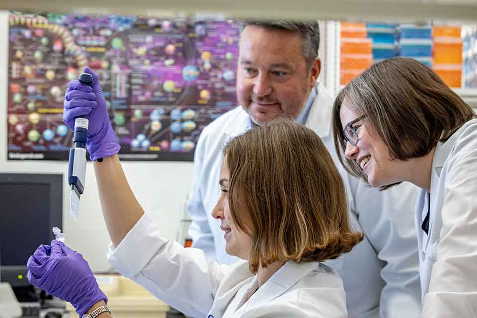 A researcher pipes a sample into a vial as two other researchers look on from behind her.