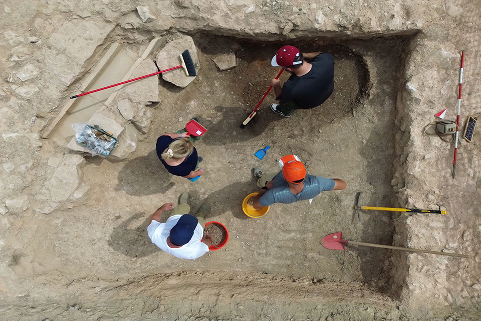 The archaeological team observes and carefully unearths underground structures at their site. The team, seen from above, works at the site with shovels, buckets and brooms.