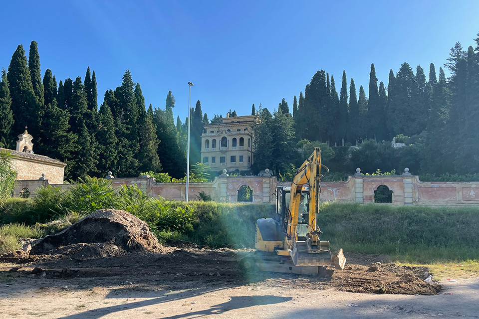 Excavator operator digs at the archeological site near Villa Fidelia in Spello, Italy. A piece of earth-moving equipment sits on a pile of dirt, with an ornate wall and home on a hillside in the background.