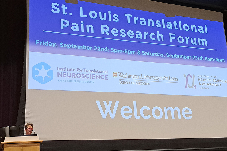 Daniela Salvemini, Ph.D., speaking at this year's Translational Pain Research Forum. A speaker stands at a podium below a large screen that reads St. Louis Translational Pain Research Forum, Friday, September 22nd: 5 p.m.-8 p.m., and Saturday, September 23rd, 8 a.m. to 4 p.m. The screen also contains logos for the Institute for Translational Neuroscience, the Washington University School of Medicine and the University of Health Sciences and Pharmacy.