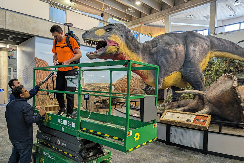 T-rex A student stands on a lift with a large piece of equipment on his back in front of one of the latex dinosaurs.