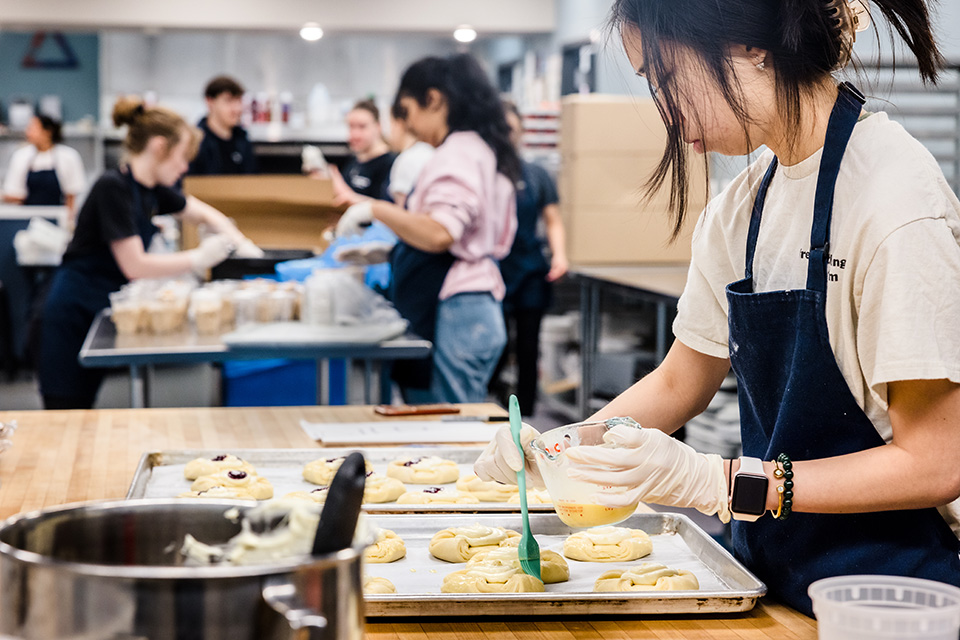 A student wearing an apron brushes melted butter from a measuring cup onto pastries on a pan while standing at a kitchen counter. Other students are at work in the background.
