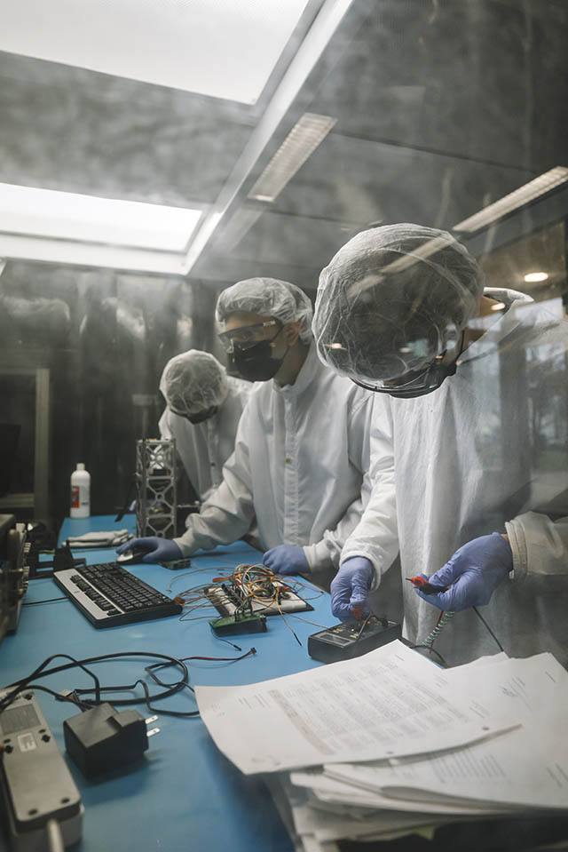 Junior and sophomore engineering students working with engineering precursors in the new Space Systems Research Lab. Three students wearing protective lab gear work at a table with various pieces of equipment and a computer.