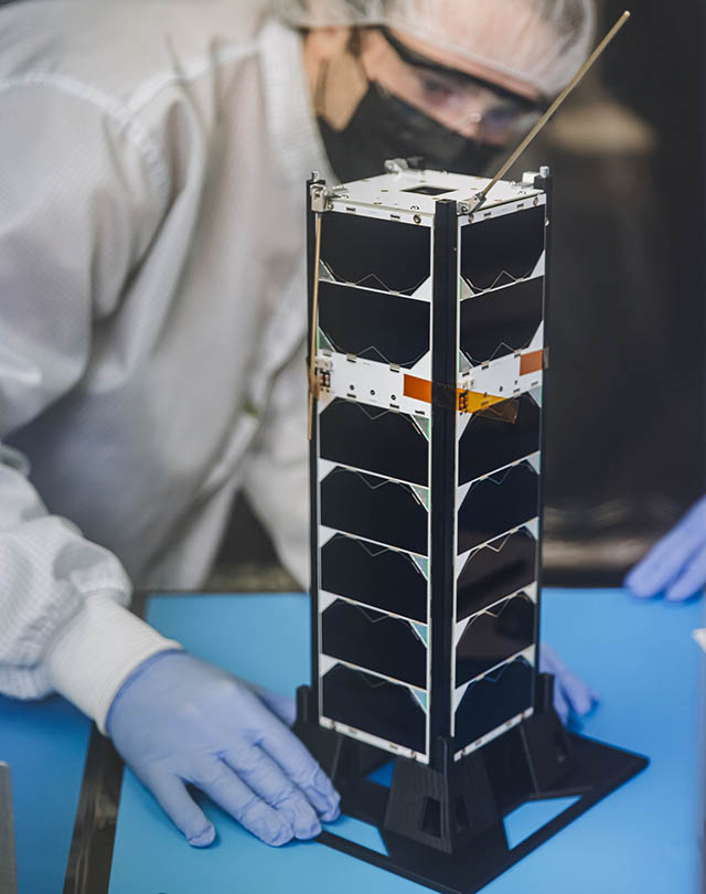 Senior engineering student, Michael Dompke, prepping DARLA for space launch. A small satellite with a height of about about 30 centimeters long and 10 centimeters on its side shaped like a metal loaf of bread sits on a table. A student wearing protective gear is seen in the background.