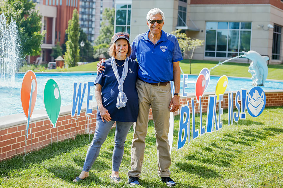 Fred Pestello with his spouse, Fran, near the Dolphin Pond during Fall Welcome Fran and Fred Pestello wear casual clothes and stand by the Dolphin Pond on SLU's campus. Colorful signs line the pond.
