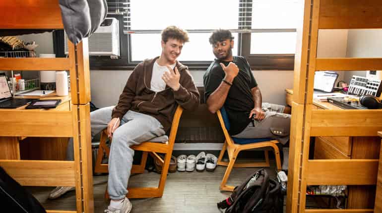 Two roomates sit and have a laugh between studies at their dorm room desks under the top level bunk beds