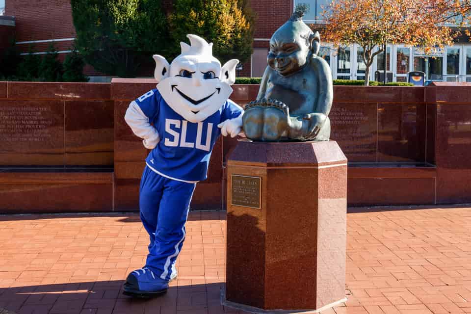 The SLU Billiken mascot poses to the left of the Billiken statue in front of Chaifetz Arena